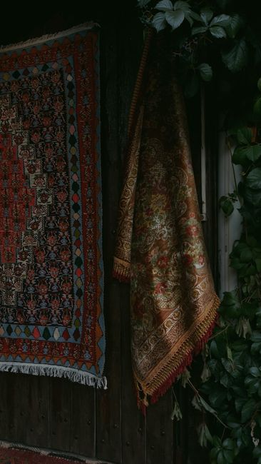 A close-up view of a traditional patterned rug with geometric designs in shades of blue, red, beige, and orange, partially draped over a polished dark wooden floor in a residential setting. The rug ha