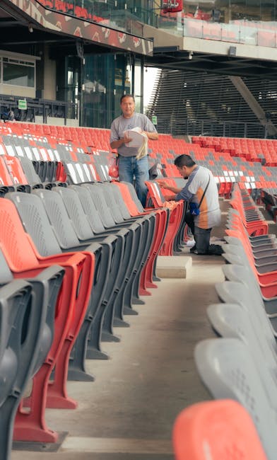 Two cleaning workers are performing surface cleaning on the red and gray stadium seats at Twickenham Stadium after an event, using cloths and cleaning solutions. The seats are made of plastic, arrange